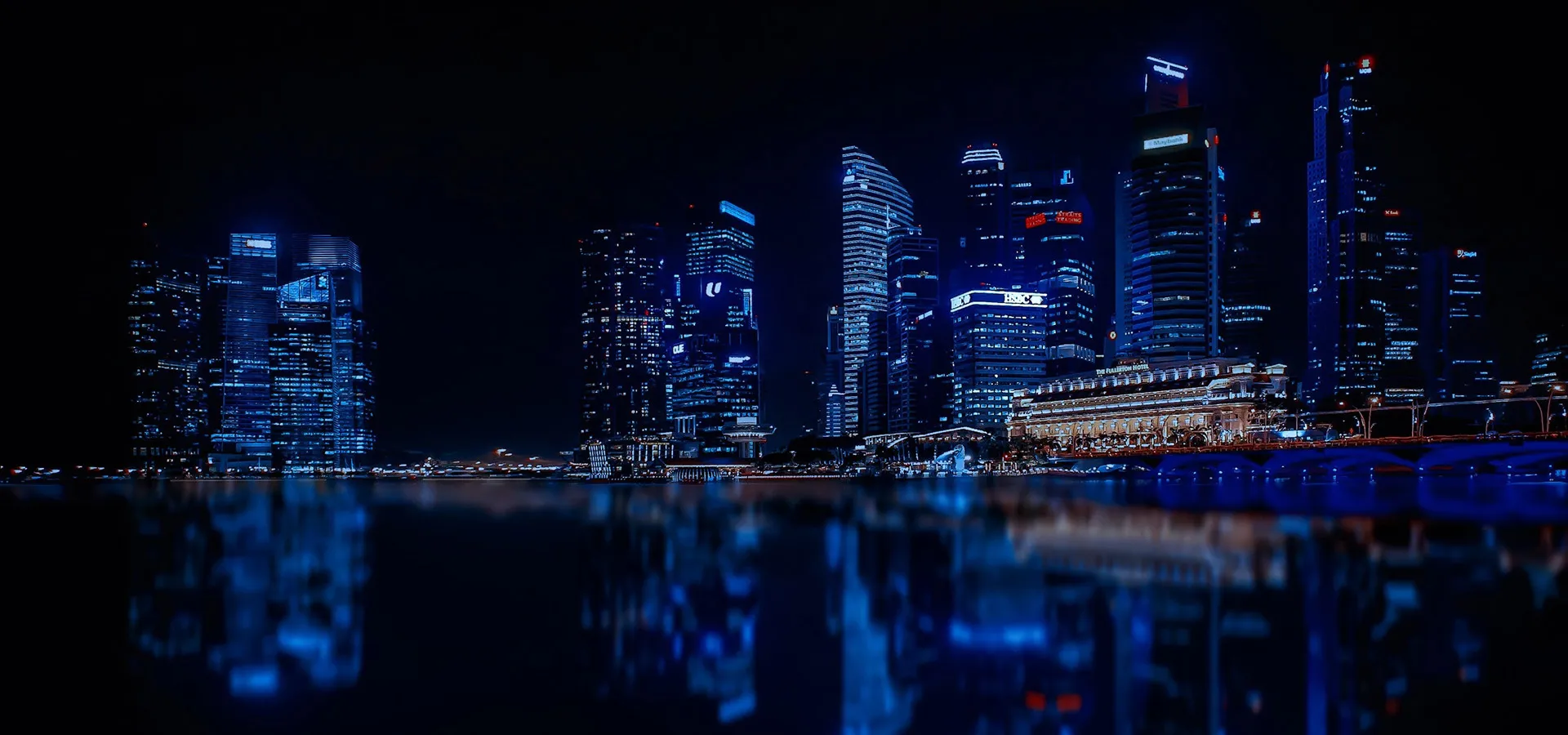Night view of a modern city skyline illuminated with vibrant blue lights, reflecting on the calm water below.