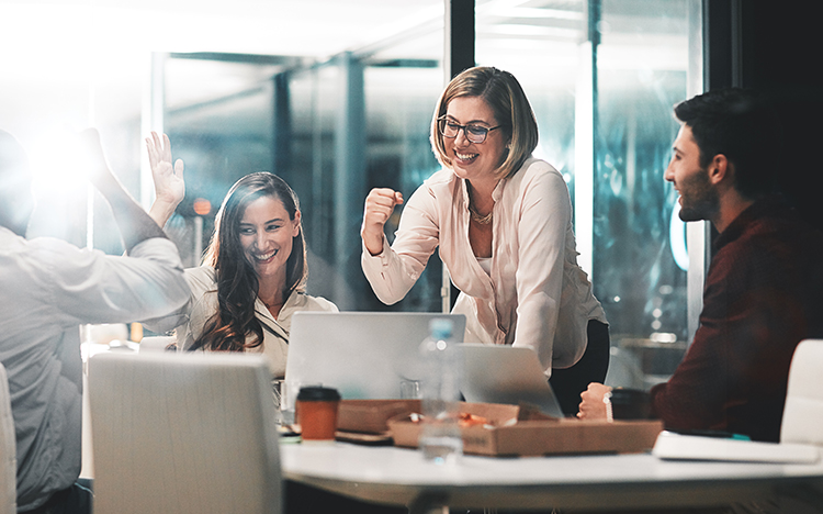Professional team celebrating successful workload automation implementation in modern office with laptops